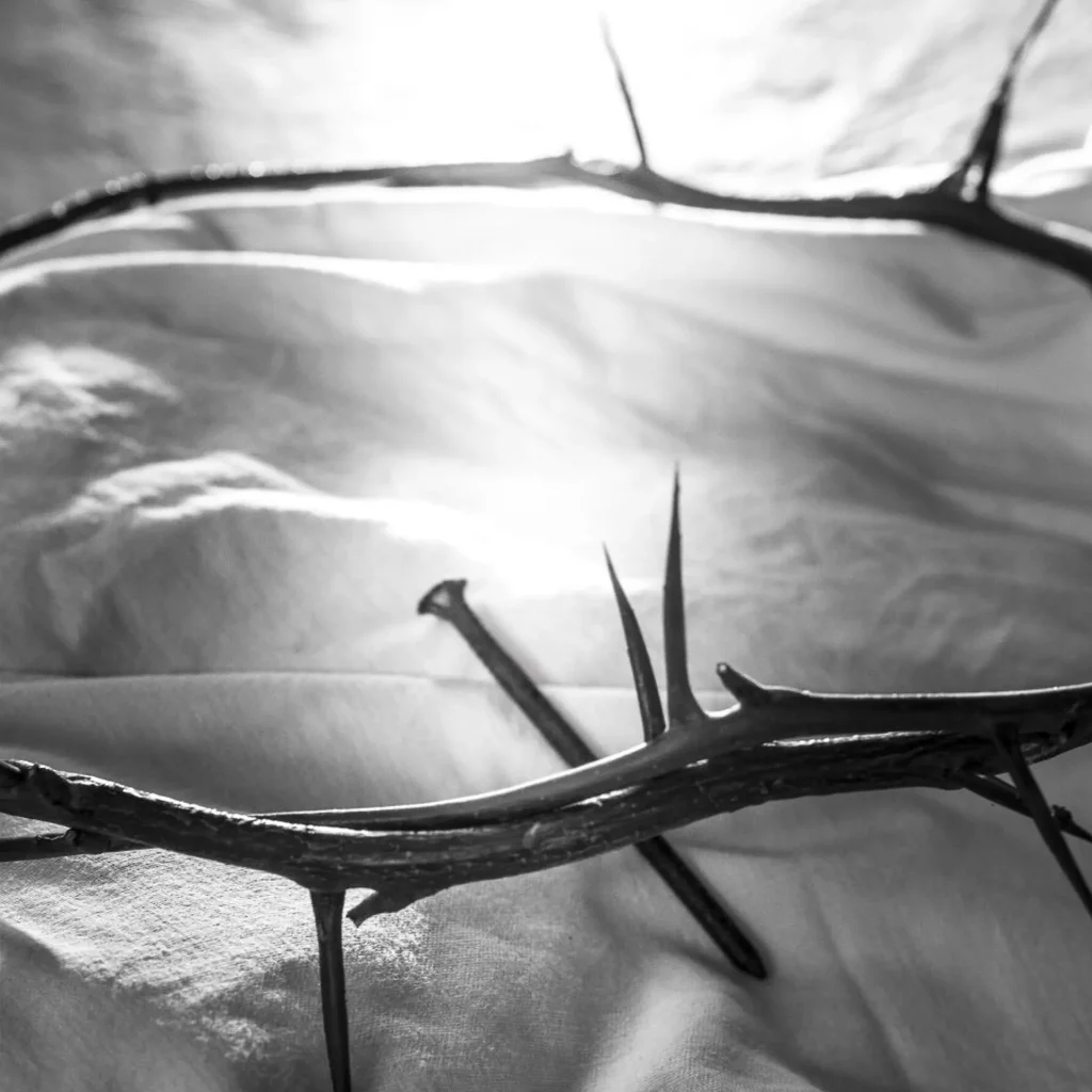 una corona de espinas sobre una tela blanca y un clavo cruzado, con la luz que viene desde atrás. Todo en blanco y negro